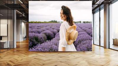 Young brunette woman standing in lavender field, wearing a bohemian white dress and a straw hat. Wall mural