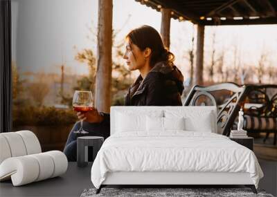 Young woman holding a glass of red wine in sunset light, outdoors at a rustic winery terrace. Wall mural