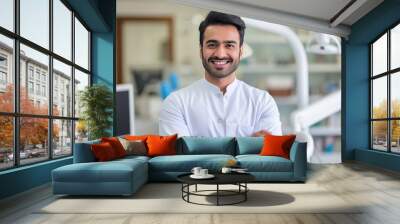 Handsome Indian male dentist smiling, standing with folded hands inside blurry modern clinic. Dental care concept Wall mural