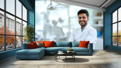 Handsome Indian male dentist smiling, standing with folded hands inside blurry modern clinic. Dental care concept Wall mural