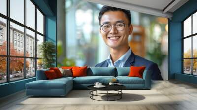 Happy young Asian businessman in stylish clothes and glasses smiling friendly against blurred modern office room background Wall mural