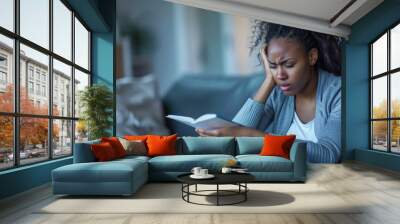 A black woman sits on a sofa. Holding his head in pain and crying with one hand as he read from an open notebook. Wall mural