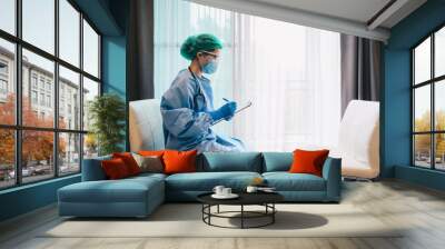 Healthcare worker wearing medical gown while filling patient information about diagnosis and prescriptions. Female therapist working alone in hospital office doing paperwork. Wall mural