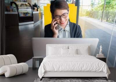 Portrait of young Asian businesswoman working on laptop while talking to someone on phone. Conceptual shot of businesswoman lifestyle while working. Wall mural