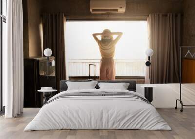 Rear view of tourist woman standing nearly window, looking to beautiful view with her luggage in hotel bedroom after check-in. Wall mural