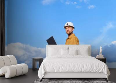 Beautiful caucasian bearded building contractor, foreman or engineer in white helmet and orange jacket using laptop at construction site at sunny day with blue cloudy sky. High quality image Wall mural