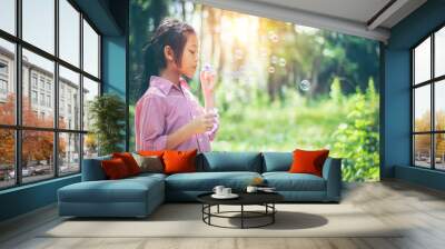 Asian girl wearing a pink shirt, being within the Park. She is currently blowing soap bubbles with fun. In the midst of the evening sun, concept of fun and freedom with copy space. Wall mural