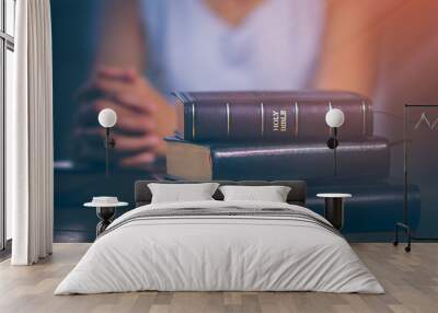 holy bible and other books stacked on a wooden table, with the hands of the young woman being prayin Wall mural