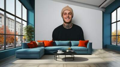 A cheerful young man with tattoos and a beanie hat smiling against a neutral background, dressed in casual black attire.
 Wall mural