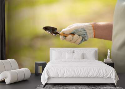 White man holds garden tool in his hand. Farmer in white cotton construction glove with blue dots holds small shovel. Spring gardening season. Autumn work in park. Green blurred background. Hobby Wall mural