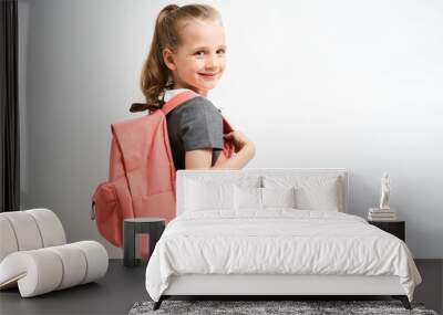 Little girl photographed against white background wearing school uniform dress isolated holding a coral backpack on both shoulders Wall mural