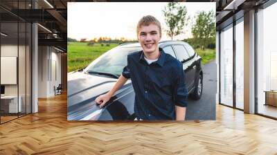 Happy young man leaning against his car next to an open field at sunset. Wall mural