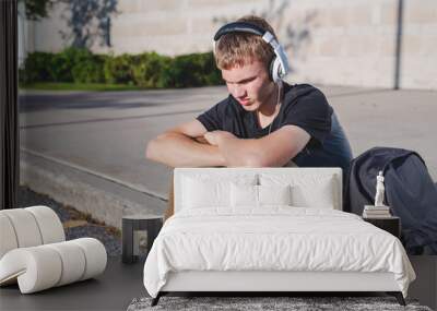 Sad teenage boy sitting on curb near school while listening to music on his headphones. Wall mural