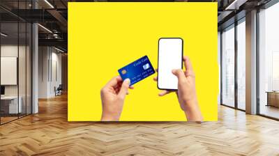 Woman hand holding blue credit card and using smart phone isolated on yellow background, front side view. Blue bank-card design mock up. mobile payment ,online shopping concept Wall mural