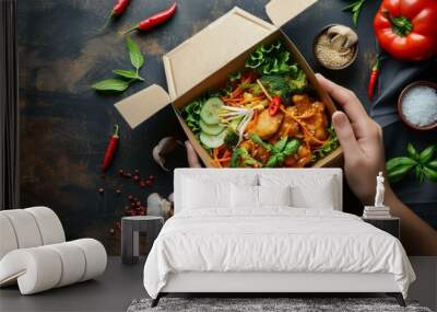 Vibrant vegetarian meal in takeout box, held with two hands, surrounded by fresh vegetables and spices, evoking healthy eating and convenience. Wall mural