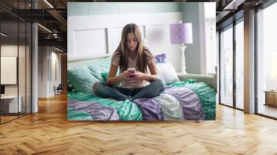 Adolescent teen girl texting on a smartphone sitting in bed at home in her bedroom Wall mural