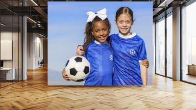 Cute, young african american and hispanic female soccer players holding a ball with a simple blue sky background Wall mural