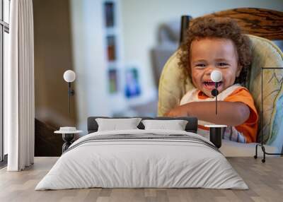 Cute Laughing diverse Child sitting in his high chair eating a meal. Happy expression as he eats a bowl of food while wearing a bib Wall mural