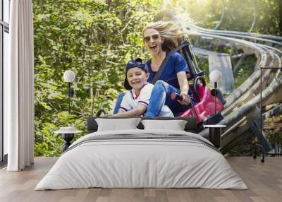 Smiling women and her boy riding downhill together on an outdoor roller coaster on a warm summer day. She has a fun expression as they enjoy a thrilling ride on a red amusement park ride Wall mural