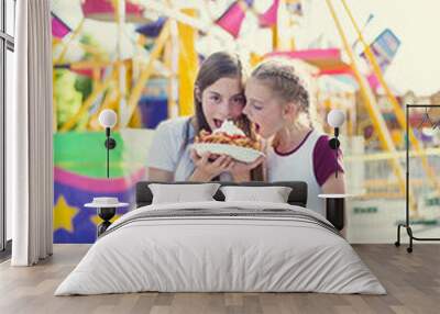 Two teen girls making a silly face while eating a funnel cake at an amusement park ride. Sticking out their tongues at pulling a funny face. Carefree and fun-loving teen girls at a carnival Wall mural