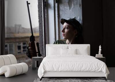 A young woman with a rifle in uniform at the window looking at the street. The woman sniper in a green suit and cap. Wall mural