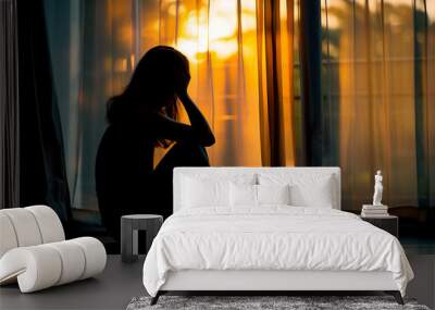 Silhouette of a pensive woman sitting by the window during sunset, with warm light casting shadows Wall mural