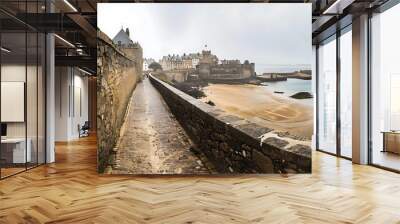 A scenic view of a coastal town with a stone pathway and sandy beach under a cloudy sky. Wall mural