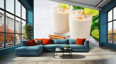 A refreshing, creamy drink garnished with cinnamon and lime, served in two glasses, on a light backdrop with tropical foliage. Wall mural