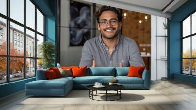 Happy Latin man wearing glasses talking to camera in modern office, sitting at desk, smiling confidently, conducting remote training Wall mural