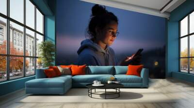 Beautiful young woman use smartphone at night. Millennial generation z female scroll through news feed on app on her phone. Cinematic film look photo of pretty woman at sunset use phone Wall mural