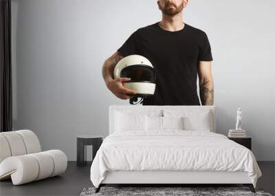 Brutal attractive bearded guy with tattooed hands poses in black blank t-shirt from premium thin cotton and holds ivory classic biker helmet, isolated on white mockup Wall mural