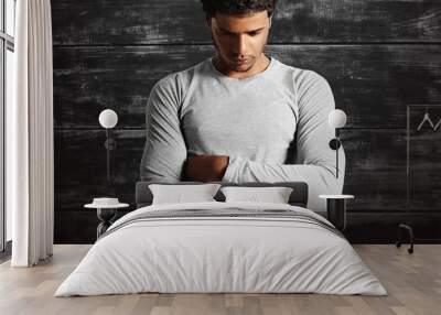 Thoughtful-looking fit dark-skinned model wearing a plain cotton light gray t-shirt with his arms crossed on the background of a black painted wooden wall. Wall mural