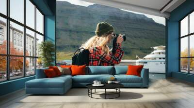 Young trndy millennial hipster, blonde woman in red shirt and green beanie makes photos of huge cruise ship entering fjords in iceland or norway. Travel blogger explores world Wall mural