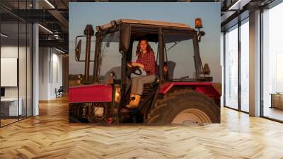 Young female farmer driving tractor in field at sundown. Wall mural