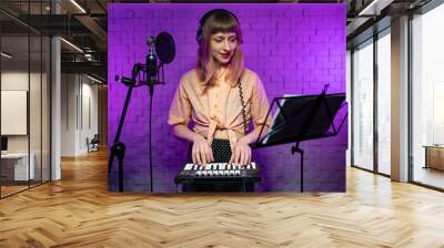 a young caucasian female learns to play a keyboard instrument on the notes, looks into the music stand next to the microphone in a recording light studio with purple light Wall mural