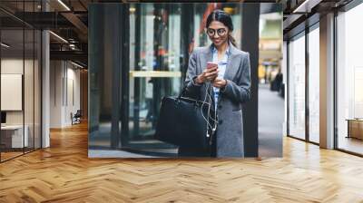  Cheerful young businesswoman texting on cellphone while walking at street Wall mural