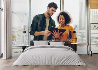 African american hipster girl together with caucasian young man reading information in book preparing for exam.Two diverse colleagues dressed in casual wear checking report standing in office Wall mural