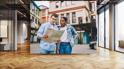 African american young woman together with caucasian friend holding map and searching right direction of route standing on street in urban setting.Trip of multicultural couple in love in new city Wall mural