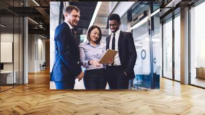 Asian cheerful woman in shirt and trousers and businessman in formal suit and African American male with toothy smile using tablet indoors Wall mural