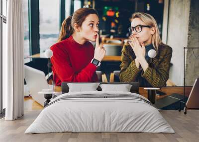 Brunette hipster blogger told secret to wondered colleague and showing sign shh during collaborating at laptop devices in coworking.Excited two best friends gossiping during studying break at netbooks Wall mural