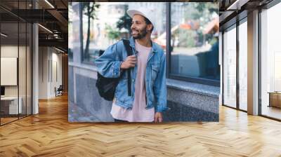 Carefree hipster guy in trendy white cap looking away and smiling during daytime sightseeing in city, happy male tourist in stylish clothing have solo vacations for exploring town during spring Wall mural