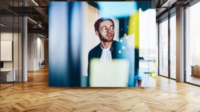 Caucasian hipster guy in optical eyewear pondering 20 years old looking away standing near bokeh window and thinking, youthful male student in trendy spectacles pensive on idea in coworking space Wall mural