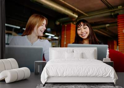 Cheerful female colleagues talking about business ideas while projecting web design via digital laptop computers, happy Asian and Caucasian women working remotely together discussing media app Wall mural