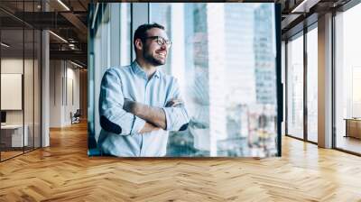 Cheerful male entrepreneur with crossed hands standing near office window view and smiling during work day in company, Caucasian successful corporate boss feeling good from wealthy lifestyle Wall mural