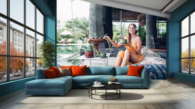 Cheerful woman with cup of tea having rest on sofa next to pool in oriental hotel Wall mural