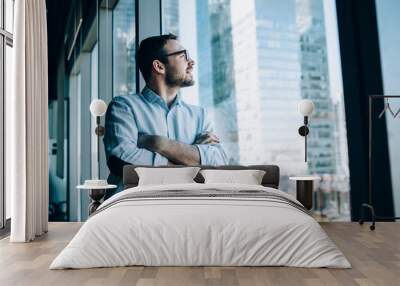 Contemplative male entrepreneur with crossed hands standing near office window view and feeling pondering during work day in company, Caucasian pensive corporate boss thoughtful looking away Wall mural