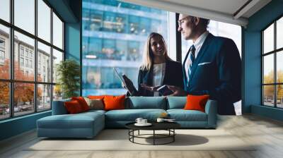 Corporate business people communicating and sharing opinions about digital tablet while standing in office interior near big window, man and woman colleagues talking and discussing work project Wall mural