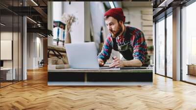 Creative male carpenter in apron browsing laptop at workshop Wall mural