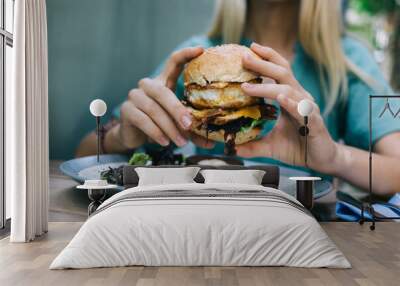 Cropped female enjoying cheat meal day for eating delicious street food - burger with mixed ingredients, woman taste sesame cheeseburger with apetito during unhealthy caloric dinner in bistro Wall mural