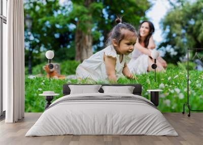 Curios kid crawling on lush green grass while mother sitting on blanket Wall mural